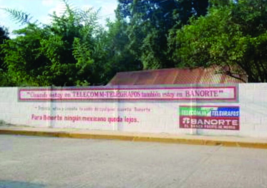 Barda pintada con el mensaje de la alianza Banorte Telecom en una calle con árboles